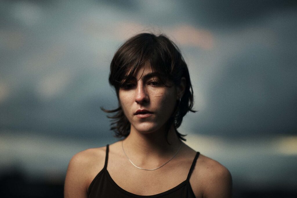 Close portrait of a person with short dark hair, wearing a black camisole and a silver necklace beneath a softly blurred cloudy sky.