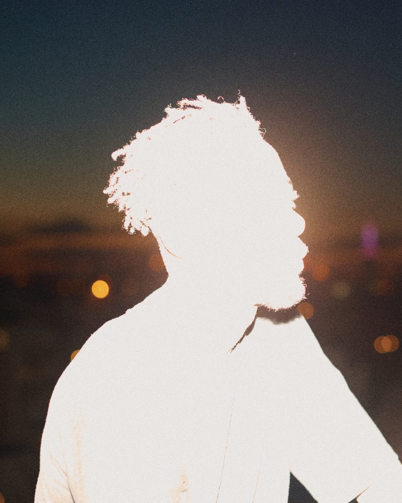 A side-profile portrait with curly hair illuminated by intense sunlight against a dark background.