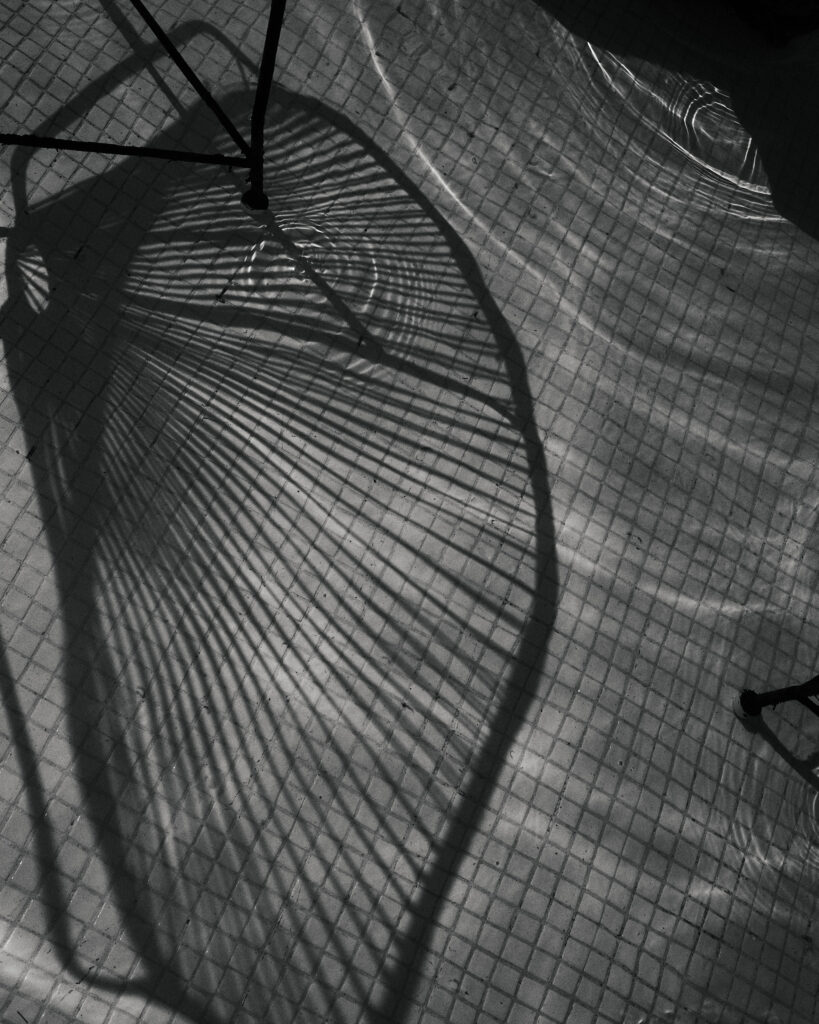 Black-and-white overhead view of a chair casting a curved, radiating shadow across a small tiled floor.
