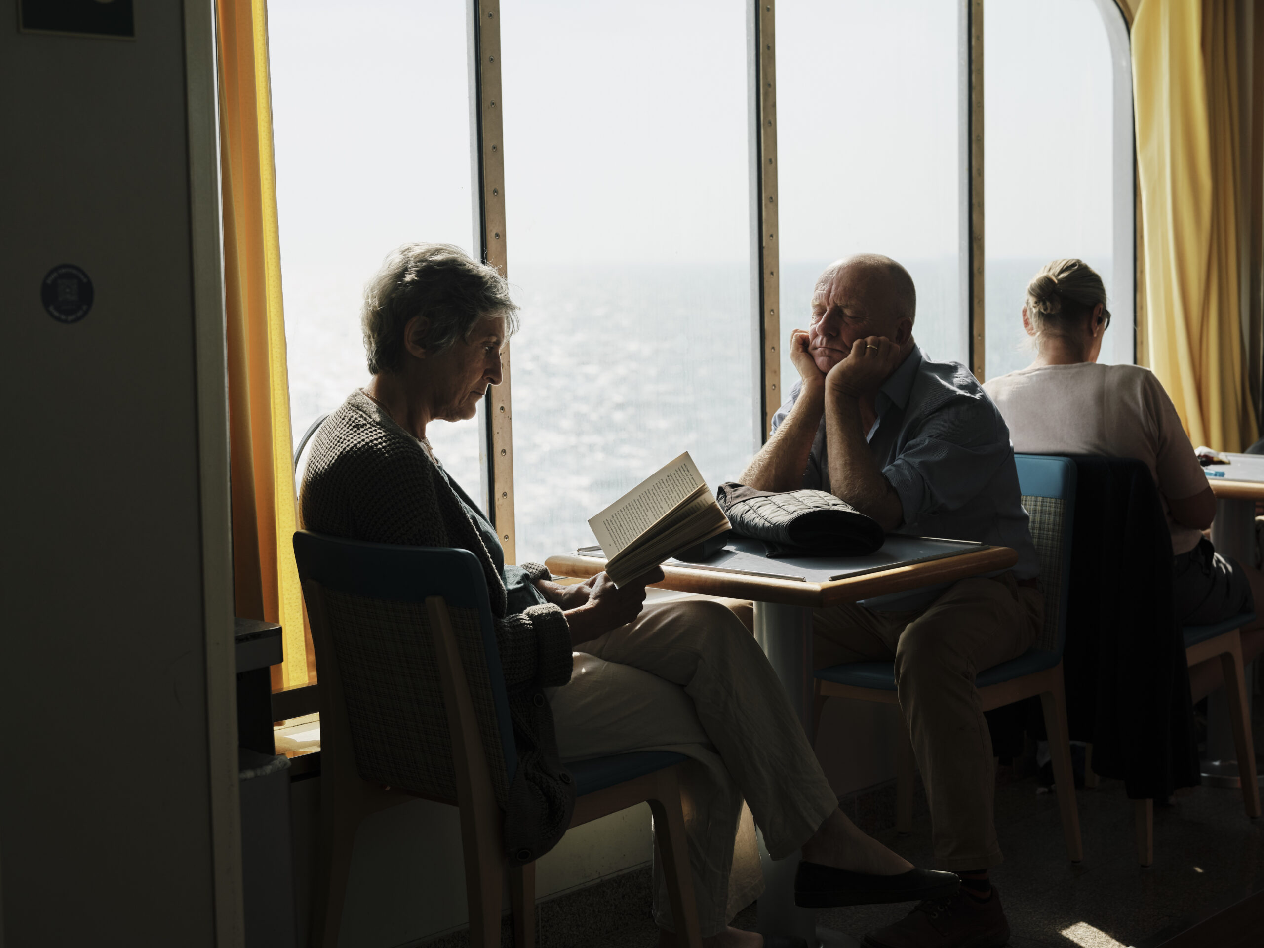 People at tables on a ferry beside a run of bright windows