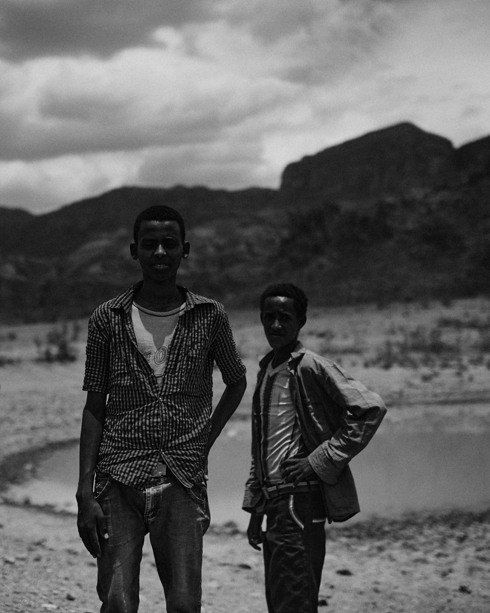 Two young men standing at the edge of a pool of water below a hillside