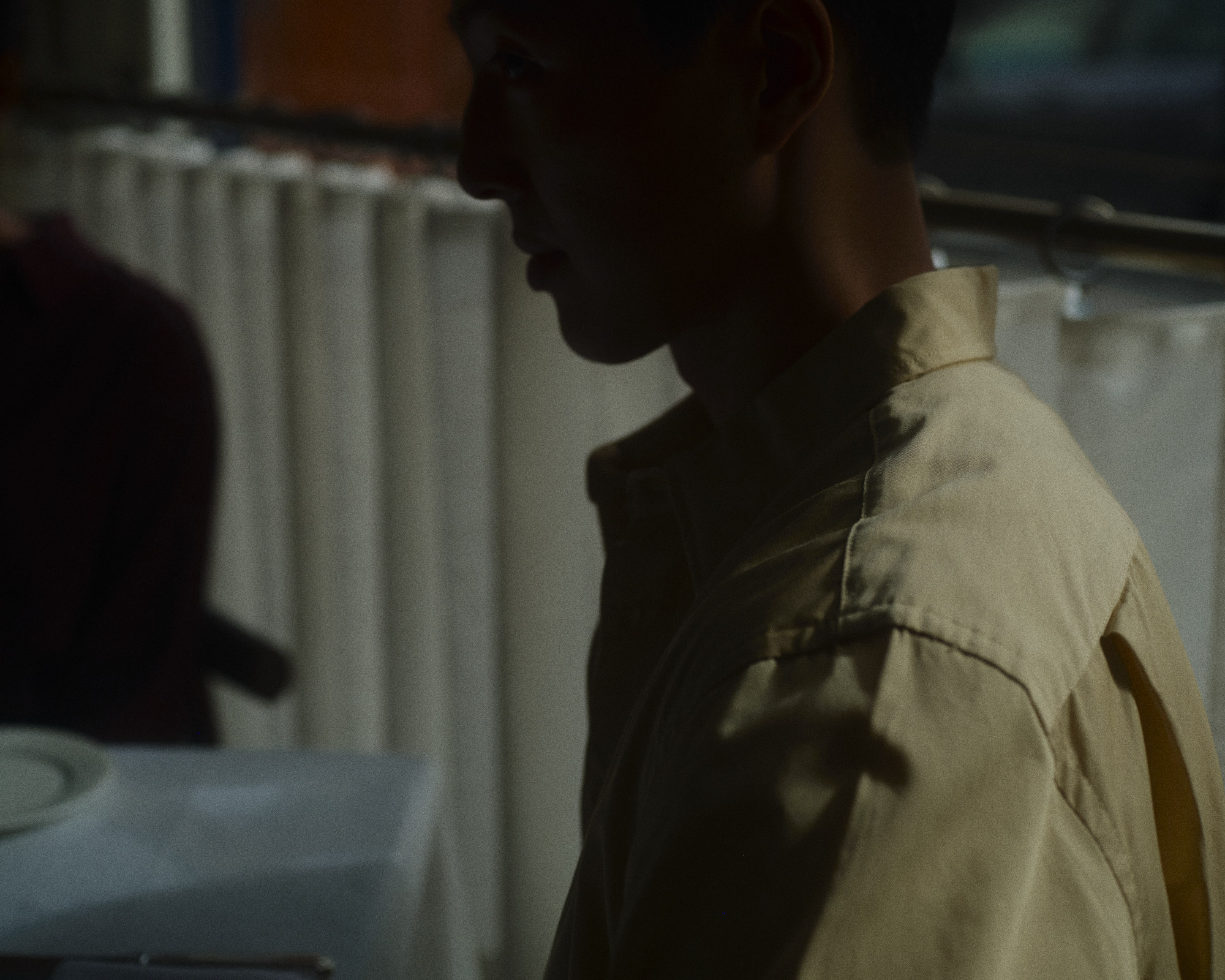 Side-profile portrait of a person in a pale collared shirt, their face in shadow against a softly lit curtain.