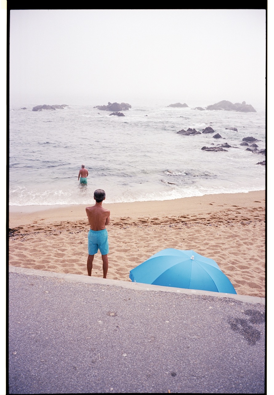Two figures in turquoise shorts facing the sea, one on the sandy beach and one wading among small waves.