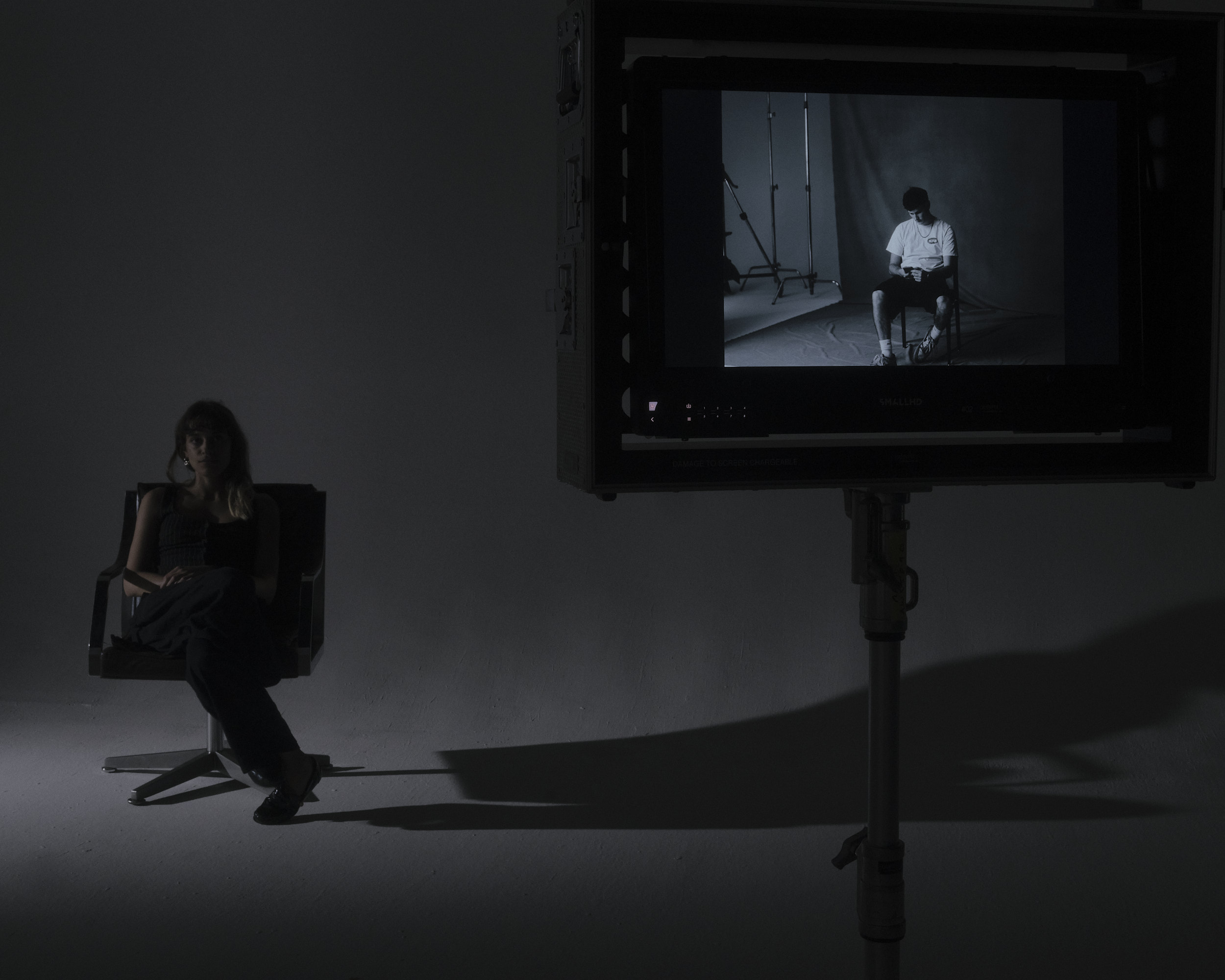 A person seated in a dark studio beside a large production monitor displaying another seated figure.